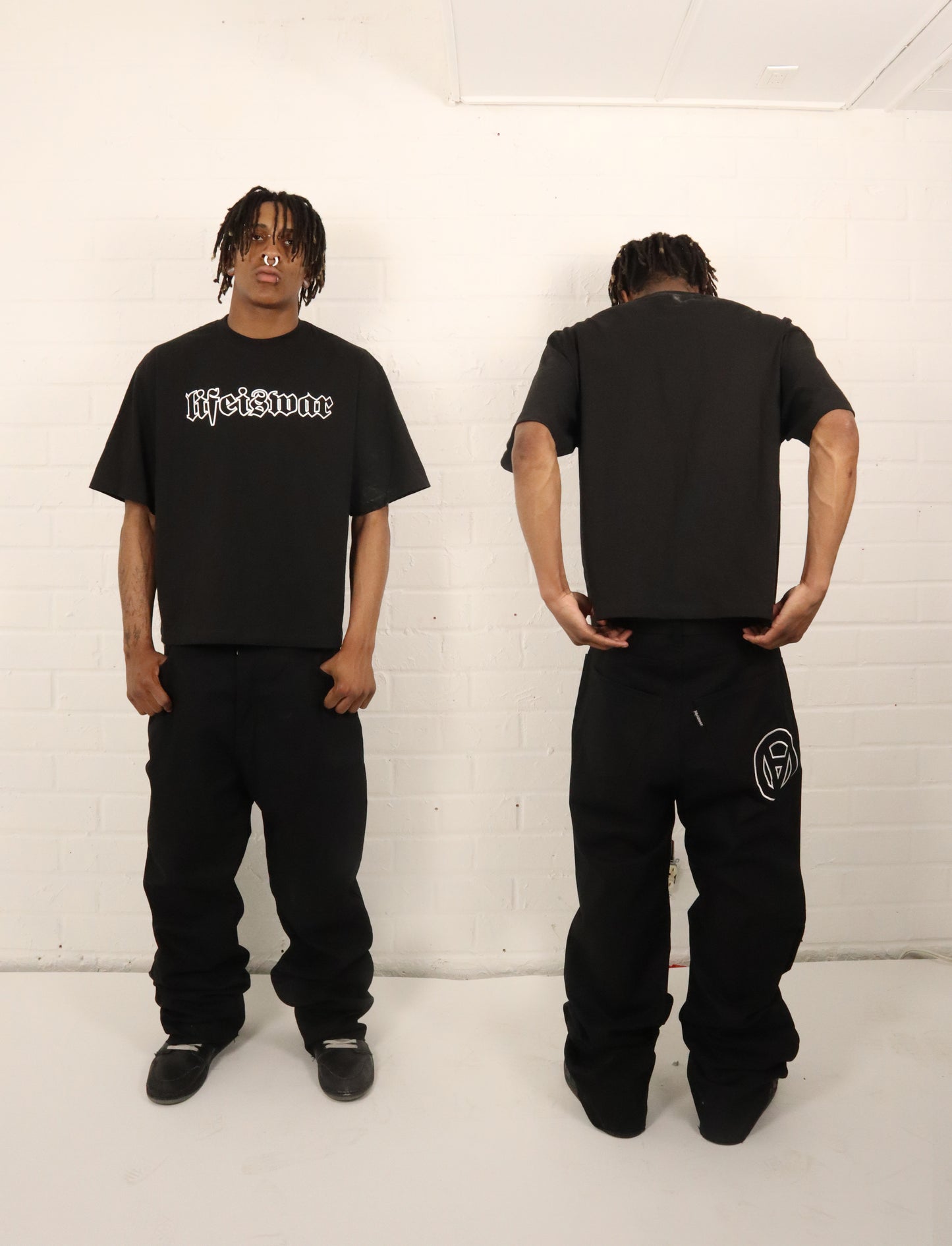 Person wearing a black t-shirt and pants with visible branding, standing against a white brick wall.