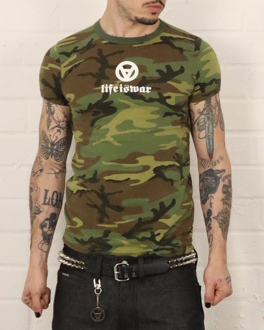 Person wearing a camouflage t-shirt with 'life is war' logo against a white brick wall.