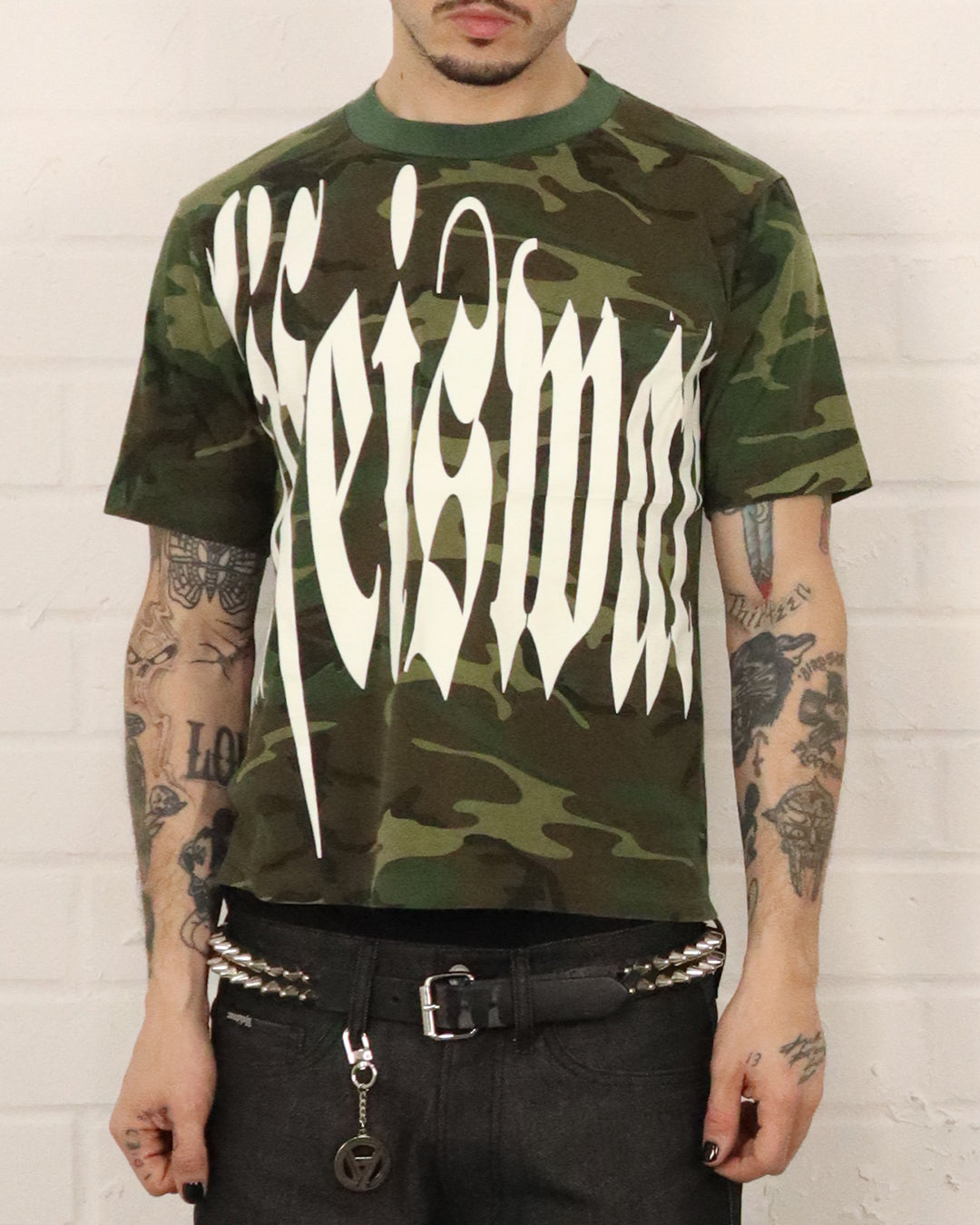 Person wearing a camouflage t-shirt with white text against a white brick wall.
