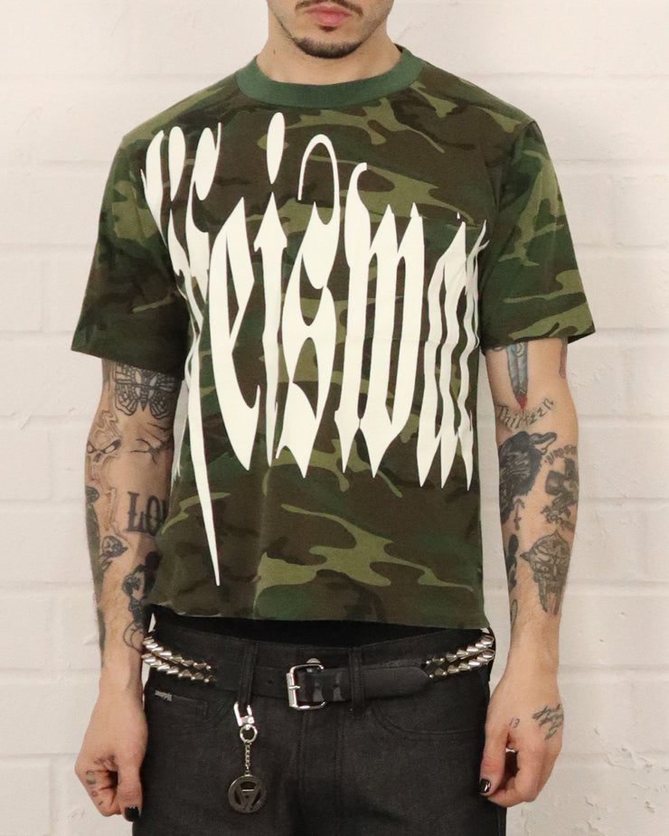 Person wearing a camouflage t-shirt with white text against a white brick wall.