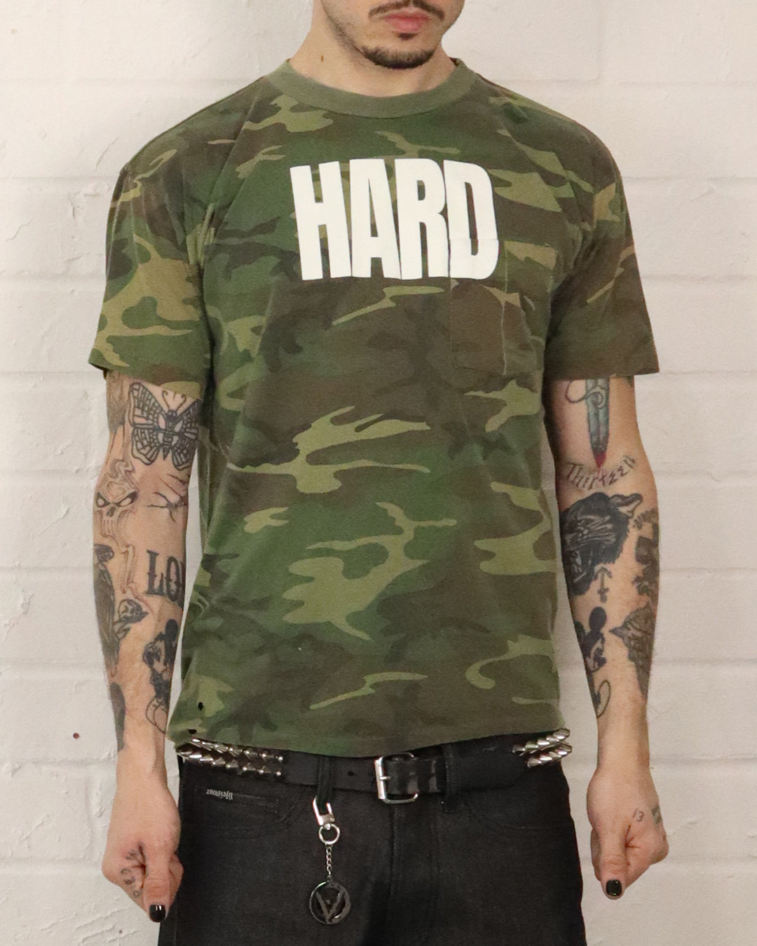 Person wearing a camouflage t-shirt with 'HARD' text against a white brick wall.