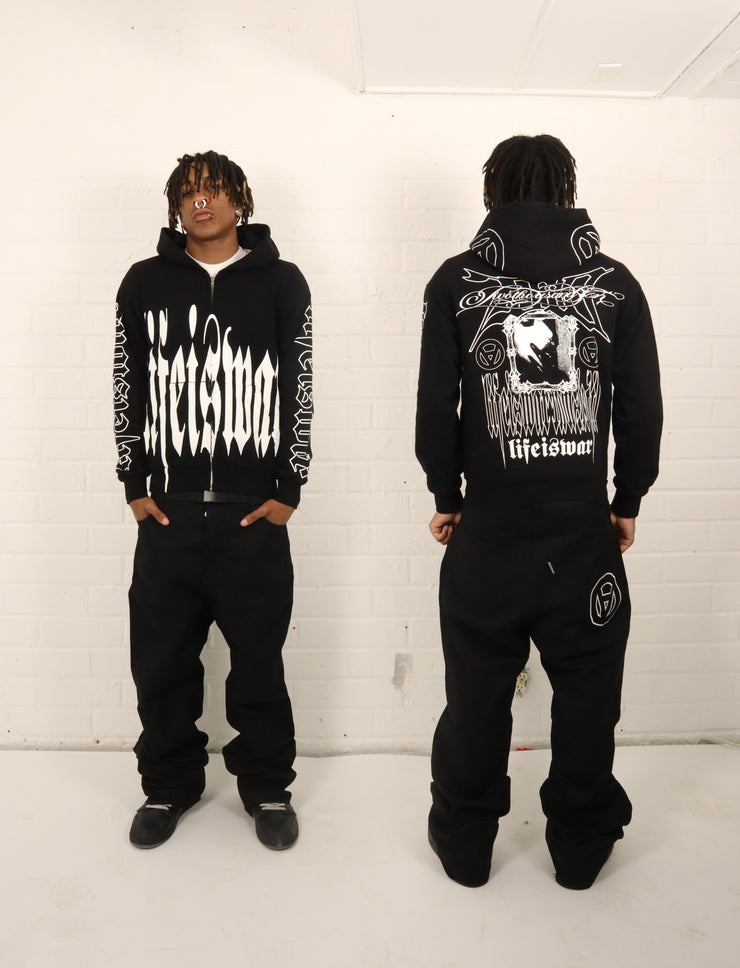 Person wearing a black hoodie with white text and graphics, standing against a white wall.