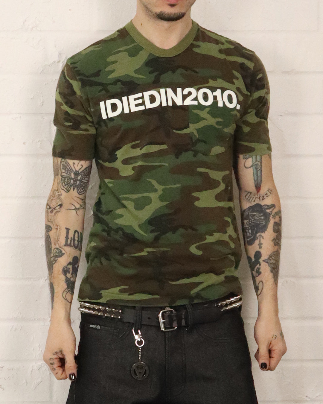 Person wearing a camouflage t-shirt with 'IDIEDIN2010' text against a white brick wall.