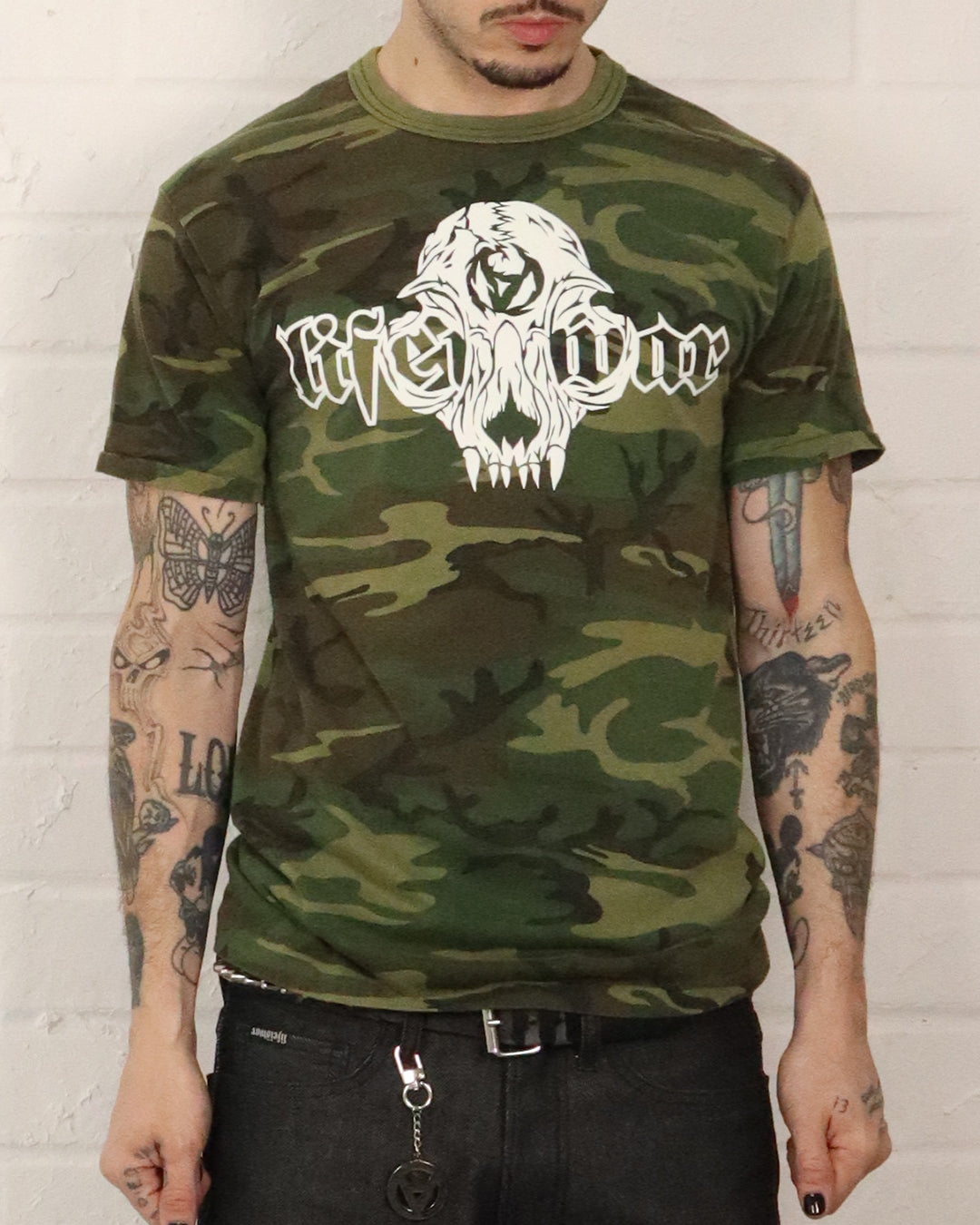 Person wearing a camouflage t-shirt with a white logo on a white brick wall background